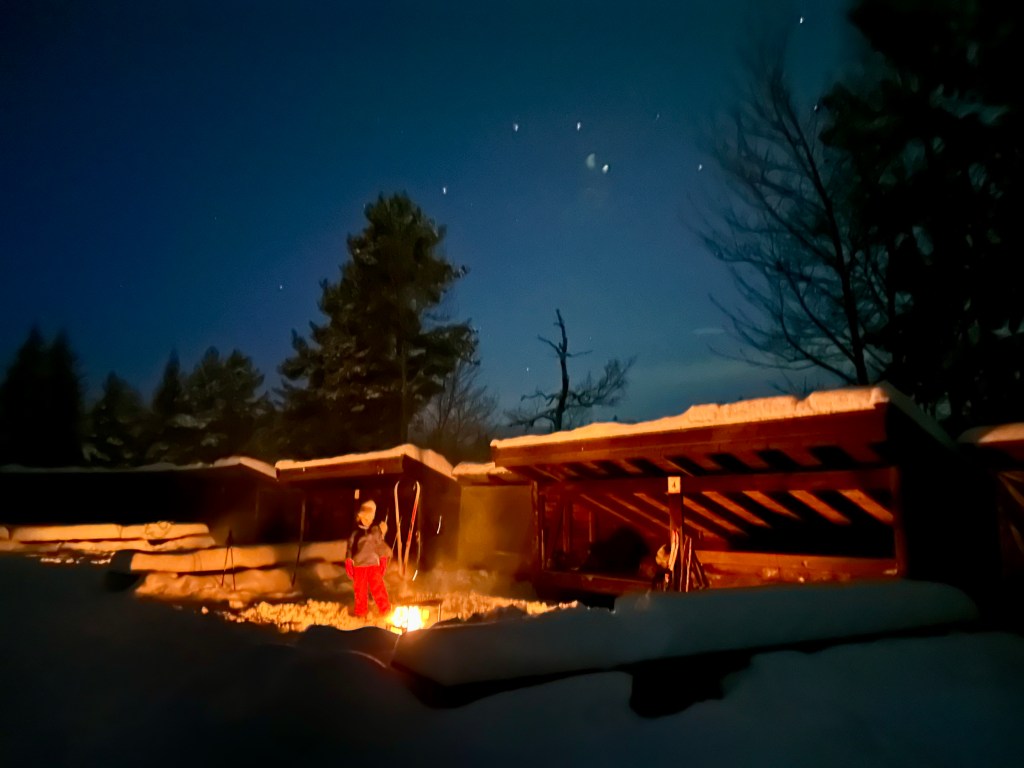 Snow-covered lean-to shelters lit by a campfire under a clear winter night sky.