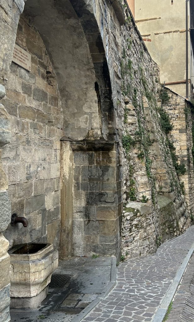 Historic stone fountain and medieval wall along a cobblestone street in Bergamo old town, Italy