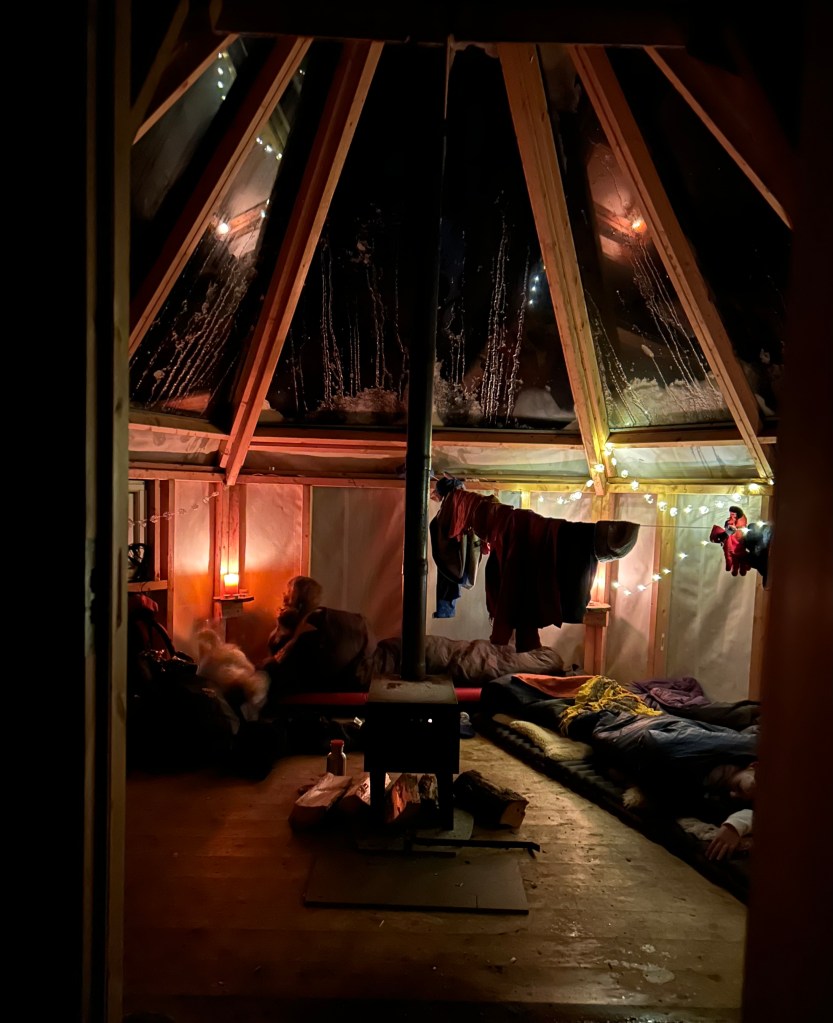 Interior of a glass-roof lavvu at night, with sleeping bags on the floor, soft light from candles, and winter gear drying inside.