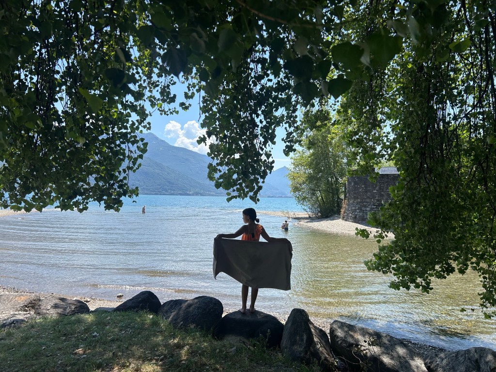 Person standing by the shore of Lake Como, Italy, surrounded by trees with mountain views and clear water