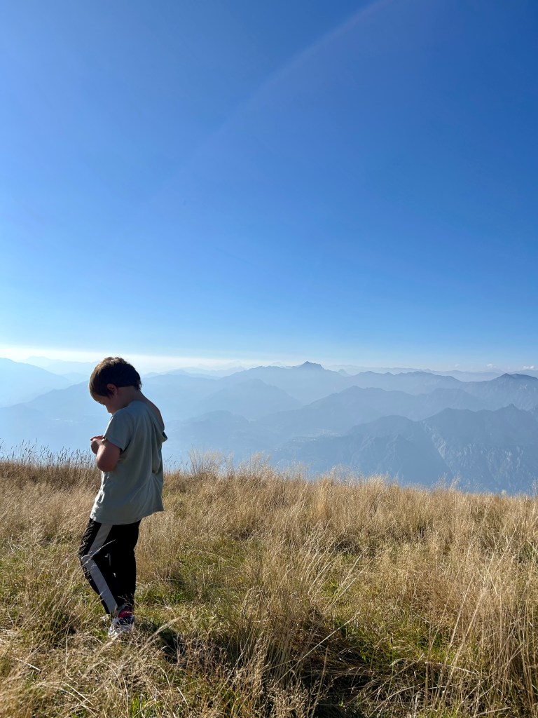 Child standing on a grassy mountain viewpoint overlooking Lake Garda, Italy, with panoramic alpine views