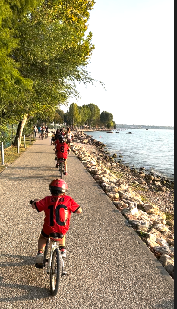 Children cycling along a scenic lakeside promenade at Lake Garda, Italy, with trees and clear blue water