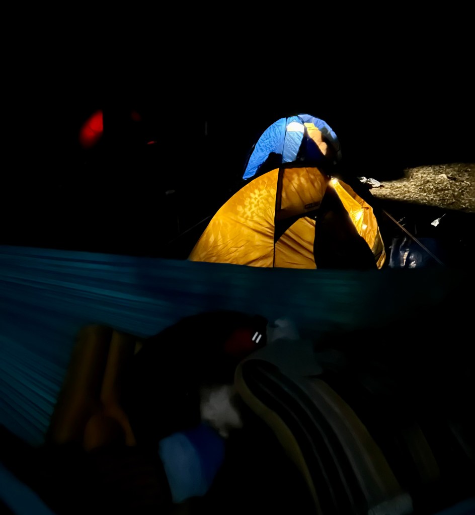 Yellow and blue tents set up in the snow at night, with a person lying in a hammock in the foreground.